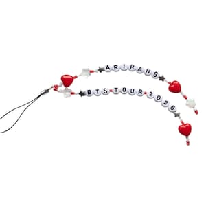 May include: A beaded phone charm with the text "ARIRANG" and "BTS TOUR 2026". The charm features red heart and star-shaped beads, along with clear and white beads. A black cord is attached for easy use.