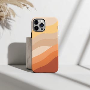 Könnte beinhalten: Eine Smartphone-Hülle mit einem wellenförmigen, geschichteten Muster in einem Verlauf aus warmen Erdtönen, darunter Nuancen von Creme, Hellgelb, Beige, Pfirsich, gebranntem Orange und Terrakotta, mit Aussparung für eine Dreifachkamera.
