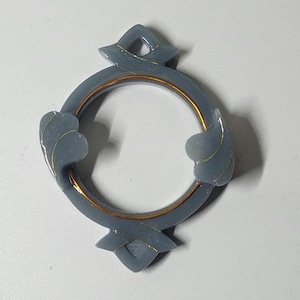 Op de afbeelding: Een grijze decoratieve ring met gouden accenten. De ring heeft een cirkelvorm met drie decoratieve elementen. De ring is gemaakt van een glad materiaal en heeft een gouden band rond de binnenste cirkel.