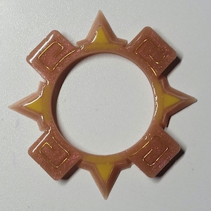 Op de afbeelding: Een cirkelvormig harsdecor met een zonnesterontwerp. Het heeft een lichtbruine basis met gele accenten en goudkleurige details. Het ontwerp bevat puntige stervormen en vierkante elementen.