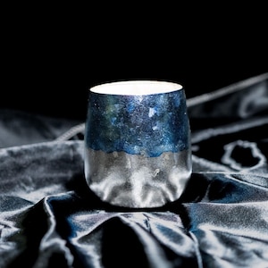 May include: A handmade ceramic tumbler with a distinctive design. The upper section of the tumbler is a deep blue with green flecks, and the lower section is metallic silver. The interior is white. It rests on a dark, textured surface.