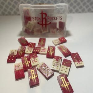 May include: A set of dominoes featuring a red and white marbled design with gold pips and the Houston Rockets logo. The pieces are scattered in front of a clear plastic storage case that displays the Houston Rockets name and team logo in red.
