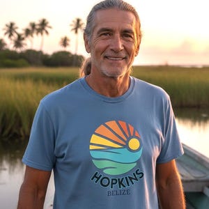 May include: A light blue t-shirt featuring a colorful circular graphic with a sun, ocean, and palm trees. The text "HOPKINS BELIZE" is printed below the graphic. The shirt is worn by a person with long gray hair, smiling in front of a natural background.