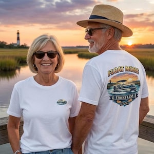 May include: Two people wearing white t-shirts with boat graphics. The shirt on the right has the text "FLOAT MORE STRESS LESS" with a boat illustration. The background shows a sunset over water with a lighthouse.