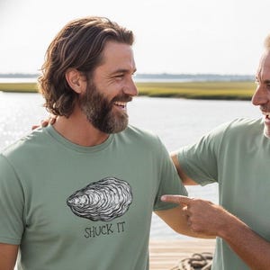 May include: Two men wearing sage green t-shirts with a black and white oyster graphic and the text "SHUCK IT". The men are smiling and pointing at each other. The background shows a body of water and a sunny sky.