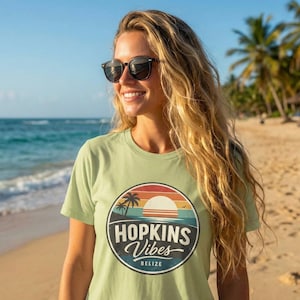 May include: A light green t-shirt with a circular graphic that reads "Hopkins Vibes Belize". The graphic features a sunset, palm trees, and ocean waves. The shirt is being worn on a beach.