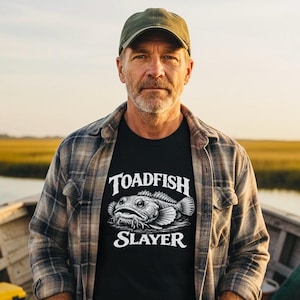 May include: A black t-shirt with the words "TOADFISH SLAYER" and a detailed illustration of a toadfish. The person is wearing a green cap and a plaid shirt. The background shows a marshy landscape.