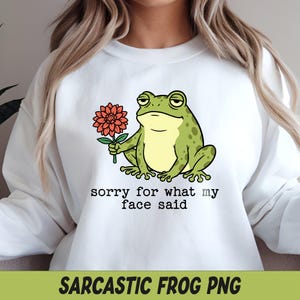May include: A white crewneck sweatshirt featuring a graphic of a green frog with a bored expression holding a red flower. Below the frog, the text reads, sorry for what my face said, in a typewriter-style font. The design is centered on the chest.