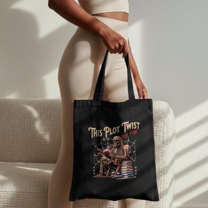 May include: Black tote bag with the text "THIS PLOT TWIST IS TO DIE FOR." The design depicts a skeleton reading a book, seated in a chair, with books and a spider web around it. The bag has black handles.