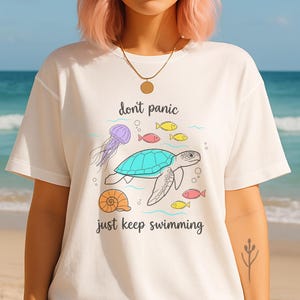 May include: White t-shirt with a sea-themed graphic. The design includes a turquoise turtle, jellyfish, fish, and a seashell. The text "don't panic just keep swimming" is also featured. The person is wearing a straw hat and sunglasses.