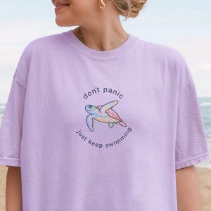 May include: A light purple t-shirt with the text "don't panic just keep swimming" and a turtle graphic. The shirt is worn with a white cap and gold earrings. The background shows a beach with sand, ocean, and palm trees.