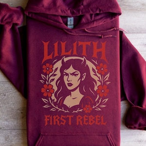 May include: Burgundy hoodie with a front graphic featuring the word "LILITH" in a stylized font above an illustration of a woman with horns, surrounded by flowers. The text "FIRST REBEL" is below the illustration. The hoodie has a hood and a front pocket.