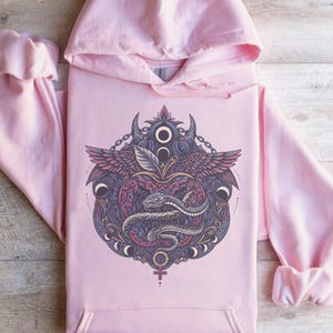 May include: A light pink hoodie with a detailed graphic on the front. The graphic features a snake, wings, and celestial elements in shades of purple, blue, and gray. The hoodie has a hood and a front pocket.