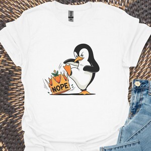 May include: White t-shirt featuring a cartoon penguin kicking a crown labeled "NOPE". The penguin is black and white with orange feet and beak. Light blue denim jeans are folded next to the shirt. The shirt is on a woven surface.