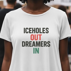 Peut inclure: T-shirt blanc avec le texte "ICEHOLES OUT DREAMERS IN" en noir, rouge et vert. Le t-shirt est porté par une personne dans une foule.
