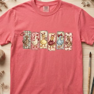 Könnte beinhalten: Ein korallenfarbenes T-Shirt mit einer Reihe von sechs Cartoon-Enten, jede mit einzigartigen Outfits und Mustern. Die Enten sind vor einem Hintergrund aus gemusterten Rechtecken platziert. Das Shirt hat einen Rundhalsausschnitt und kurze Ärmel.