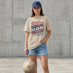 May include: A beige t-shirt with the text "USA WORLD CUP 2026" in red, white, and blue, worn with denim shorts and a navy baseball cap. A vintage football is on the ground in front of the person.