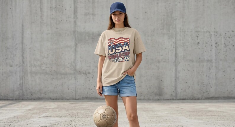 May include: A beige t-shirt with the text "USA WORLD CUP 2026" in red, white, and blue, worn with denim shorts and a navy baseball cap. A vintage football is on the ground in front of the person.
