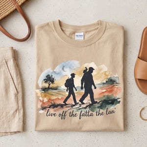 以下が含まれることがあります： 「live off the fatta the lan'」という文字と、風景の中を歩く2人の人物の水彩画風グラフィックが描かれたベージュのクルーネックTシャツ。ベージュのリネンショートパンツ、編み込みのストローバッグ、茶色のスライドサンダル、ゴールドのジュエリーが添えられています。