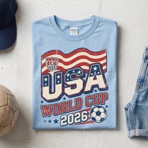 May include: A light blue t-shirt with a USA World Cup 2026 graphic, a navy blue baseball cap, a vintage football, and denim shorts. The shirt features a distressed American flag design and football graphic.
