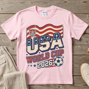 May include: A pink t-shirt with a USA World Cup 2026 graphic, alongside beige trousers, a baseball cap, trainers, and a green bandana. The items are displayed on a wooden surface, creating a casual, sporty aesthetic.