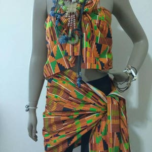 May include: A colorful orange, green, and black patterned fabric wrap is worn as a top and skirt. The fabric is tied at the waist and the top is strapless. The model is wearing a variety of necklaces with beads and charms.