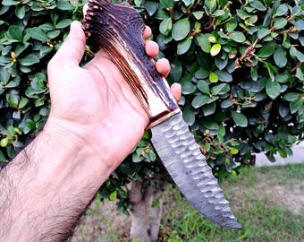 May include: A handcrafted knife with a textured blade and a handle made from a dark brown antler. The blade has a unique pattern and a curved edge. The handle has a natural, rustic appearance.