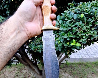 May include: A handcrafted knife with a light-colored wooden handle and a curved, patterned blade. The blade has a dark, textured appearance. The knife is held against a backdrop of green foliage and a tree.