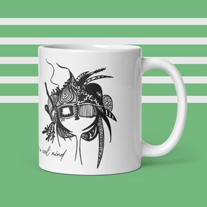 May include: White ceramic mug with a black and white abstract face design. The mug has a curved handle and the text "a cool mind". The design features intricate patterns. The background is a green and white striped pattern.