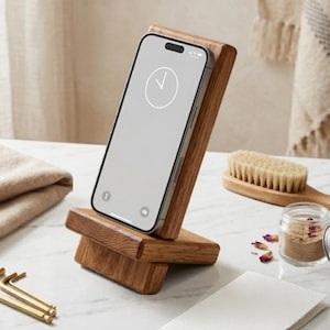 Könnte beinhalten: Handyhalter aus Holz mit einem Telefon, das eine Uhr anzeigt. Der Ständer ist aus hellbraunem Holz gefertigt und hat ein schlichtes, modernes Design. Weitere Gegenstände im Bild sind eine Bürste, ein kleines Glas und ein Stapel gefalteten Stoffes.