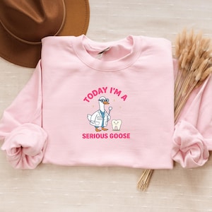 Puede incluir: Una sudadera rosa claro con el texto "TODAY I'M A SERIOUS GOOSE". El gráfico presenta un ganso de dibujos animados vestido de dentista, sosteniendo un espejo dental junto a un diente.