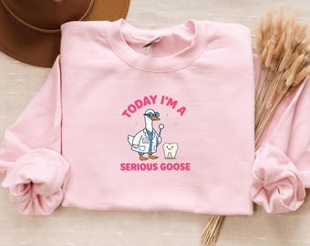 Op de afbeelding: Een lichtroze sweatshirt met de tekst "TODAY I'M A SERIOUS GOOSE". De afbeelding toont een cartoon gans gekleed als tandarts, met een tandartsspiegel naast een tand.