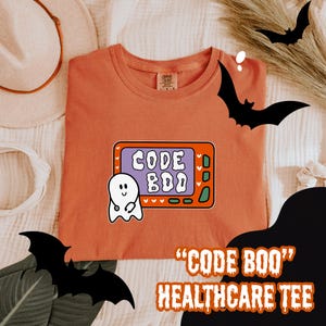 May include: Orange t-shirt with a graphic that says "CODE BOO" in a retro style, featuring a ghost and a television screen design. The text "HEALTHCARE TEE" is below the graphic. Halloween-themed apparel.