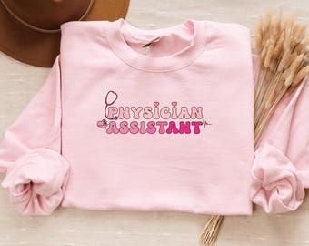 May include: A pale pink crew neck sweatshirt featuring the text "Physician Assistant" in a retro font, with a stethoscope graphic. The text is in shades of pink. The sweatshirt is a casual style.