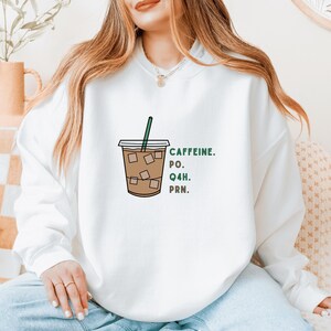 Op de afbeelding: Witte sweatshirt met een afbeelding van een ijskoffie en een groen rietje. De tekst "CAFFEINE. PO. Q4H. PRN." is in het groen gedrukt. Het ontwerp is casual en geschikt voor dagelijks gebruik. De sweatshirt is comfortabel.