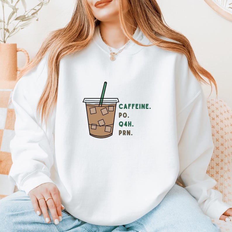 Op de afbeelding: Witte sweatshirt met een afbeelding van een ijskoffie en een groen rietje. De tekst "CAFFEINE. PO. Q4H. PRN." is in het groen gedrukt. Het ontwerp is casual en geschikt voor dagelijks gebruik. De sweatshirt is comfortabel.