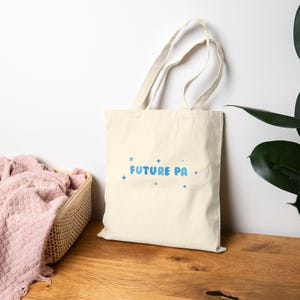 May include: Cream-colored tote bag with the text "FUTURE PA" in blue bubble letters and blue star accents. The bag has long handles and is made of a sturdy fabric, suitable for carrying books or groceries.