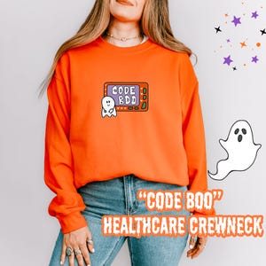 May include: An orange crewneck sweatshirt with the text "CODE BOO" and "HEALTHCARE CREWNECK" in a Halloween-themed font. The sweatshirt features a cartoon ghost and a graphic that says "CODE BDD".