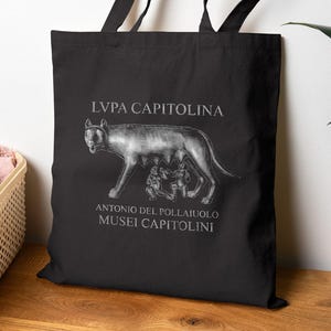 May include: Black tote bag featuring a detailed illustration of the Capitoline Wolf with Romulus and Remus. The text "LUPA CAPITOLINA" is above the image, and "ANTONIO DEL POLLAIUOLO MUSEI CAPITOLINI" is below. The bag has black handles.
