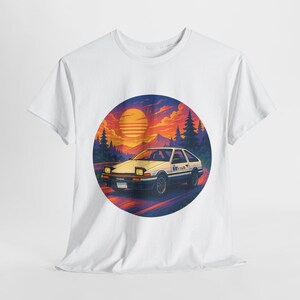 Pode incluir: T-shirt branco com um gráfico de um carro branco numa estrada, com um pôr do sol ao fundo. O carro tem detalhes pretos e texto japonês. O pôr do sol apresenta cores laranja, amarelo e azul, com árvores e montanhas no fundo.