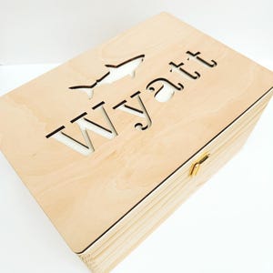 May include: A rectangular light-colored wooden box with a hinged lid. The top of the lid features a laser-cut design consisting of the name Wyatt in a sans-serif font and a stylized fish silhouette positioned above the text. A small gold metal latch.