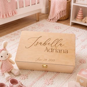 May include: A rectangular light-colored wooden keepsake box featuring the engraved name Isabella Adriana and the date June 24, 2025. The box has a smooth finish, a hinged lid, and a small gold-toned metal latch centered on the front edge.