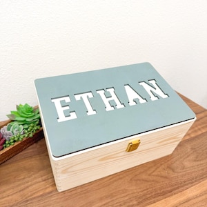 May include: Wooden keepsake box with a light blue lid featuring the name "ETHAN" cut out in white letters. The box has a natural wood finish, a gold-colored clasp, and is sitting on a wooden surface.