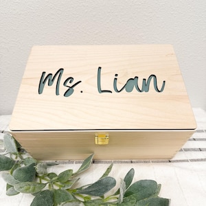 May include: A rectangular light-colored wooden box with a hinged lid featuring the laser-cut text "Ms. Lian" on the top surface. The box includes a small gold-colored metal latch on the front center to secure the lid closed.