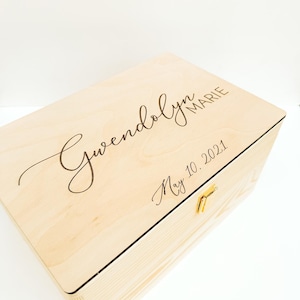 May include: A light-colored wooden box with a hinged lid and a gold-colored clasp. The lid is engraved with the name "Gwendolyn Marie" and the date "May 10, 2021" in a delicate script. The box is made of light-colored wood.