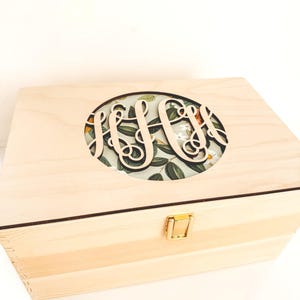 May include: A rectangular light-coloured wooden box featuring a circular cutout on the lid. The cutout displays a floral pattern with green leaves and white flowers, overlaid with a dark, script-style monogram. A small gold-toned metal latch is centred.