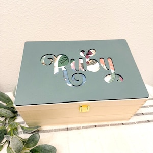 May include: A rectangular wooden box with a matte sage green lid featuring the name Ruby cut out in a cursive font. The cutout reveals a floral pattern underneath with pink, green, and blue tones. The box has a small gold metal latch on the front.