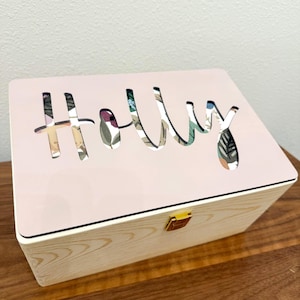 May include: A rectangular wooden box with a light-colored wood grain base and a smooth, pale pink lid. The lid features a cutout of the name Holly, revealing a colorful floral and leaf pattern underneath. A small gold-colored metal latch is centered.