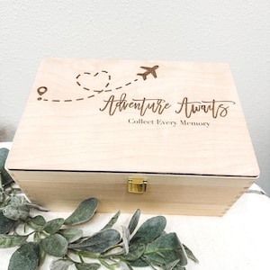 May include: A light-colored wooden memory box with a gold clasp. The lid is engraved with a dotted line, airplane, and the words "Adventure Awaits, Collect Every Memory."