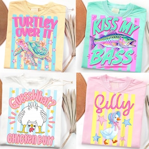 May include: Four colorful t-shirts with playful graphics. One shirt is yellow with a turtle and the text "TURTLEY OVER IT". Another is mint green with a fish and the text "KISS MY BASS". A white shirt has a chicken and the text "GUESS WHAT? CHICKEN BUTT". The last is pink with a duck and the text "SILLY".
